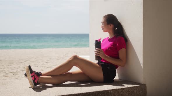 Sportive Woman with Bottle of Water Relaxing After Workout on Beach