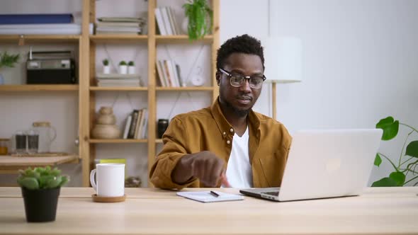 African Man with Glasses Works on a Laptop and Writes in a Notepad on Table Spbas