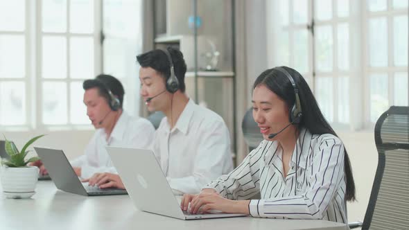 Three Asian Call Centre Agents Wearing Headsets And Typing On Computer While Speaking To Customers