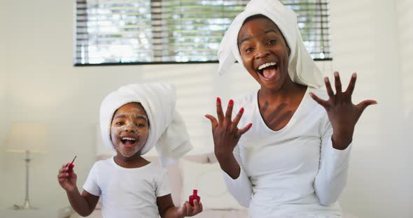 Happy african american mother and daughter sitting on bed and showing painted nails to camera