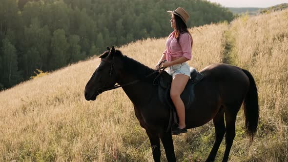 Beautiful Woman in Hat Sitting on Horseback Among the Mountains. Smiling Woman on Brown Horse Posing