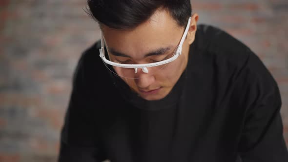 Man with Face Shield Looks Down Against Blurry Brick Wall