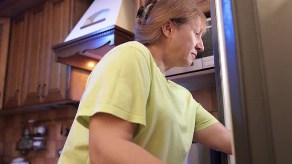 Middle-aged woman opens refrigerator at night and drinks milk from a plastic bottle.