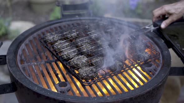 Man puts a grilling basket full of browning kofta on the grill. Camera moves right to left.