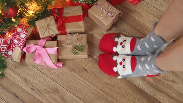 Feet in Funny Christmas Socks Santa Face Wooden Floor Under Fir Tree Gift Boxes