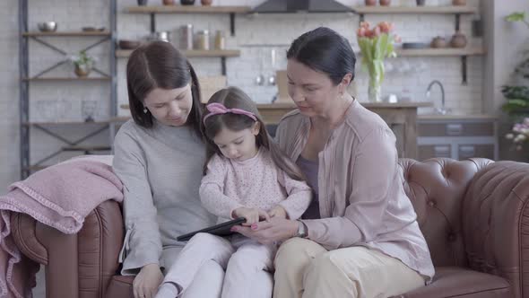 Grandmother, Mother and Little Daughter Sitting Together on the Sofa in Modern Studio Apartment. The