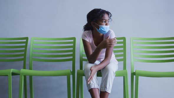 African american girl lowering her face mask while sitting on a chair at hospital
