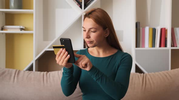 Young Woman Customer Holding Credit Card and Smartphone Sitting on Couch at Home. 