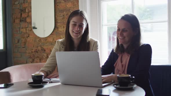 Two smiling caucasian businesswomen sitting at table with coffee, using laptop and talking,