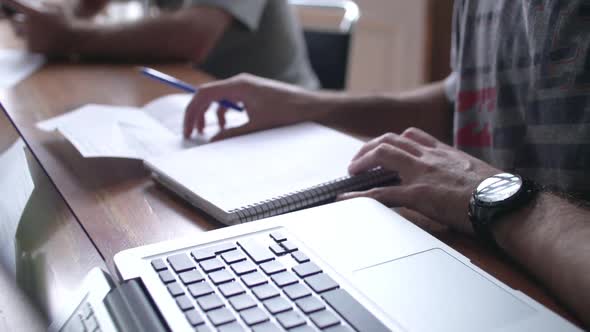 Close-up of man writing in notebook with laptop computer nearby