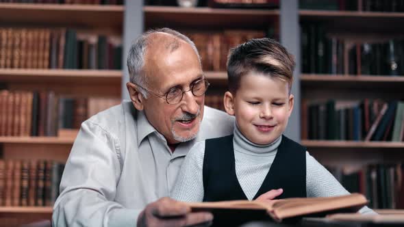 Happy 70s Elderly Grandfather and Little Schooler Grandson Reading Book at Library