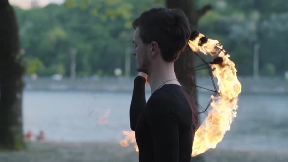 Portrait of Young Professional Man Performing a Show with Fire Fan Standing on Riverbank in Front of