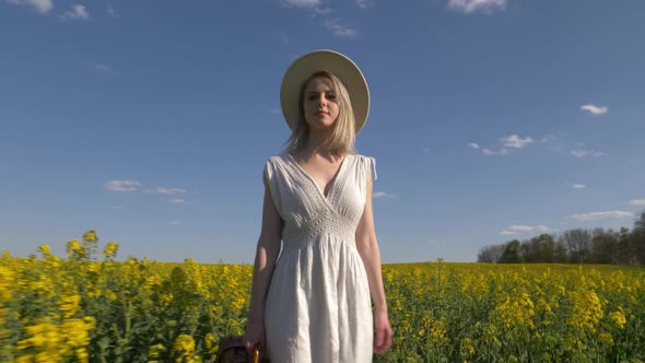 Blonde woman in white dress with suitcase in rapeseed field in spring time