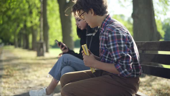 Teenage Girl Surfing Internet on Phone in Sunshine in Park As Boy Approaching Sitting Down on Bench