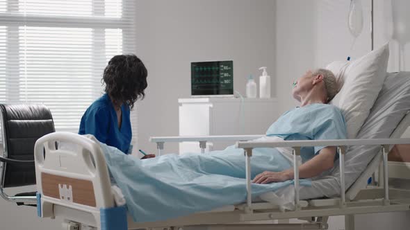A Black Woman Cardiologist Doctor is Talking to an Old Man Patient Lying on a Hospital Bed
