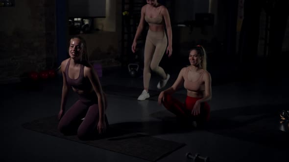 Wide Shot Joyful Young Women Start Training in Gym Looking Away Sitting Down on Exercise Mats