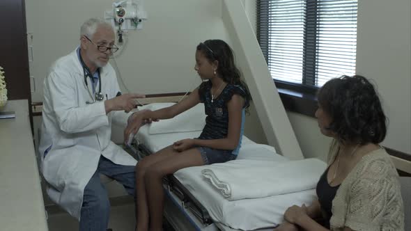 Doctor examines the wrist of a young girl. With mother in room.