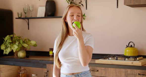 Young blonde woman is throwing up green apple and eating it in the kitchen.