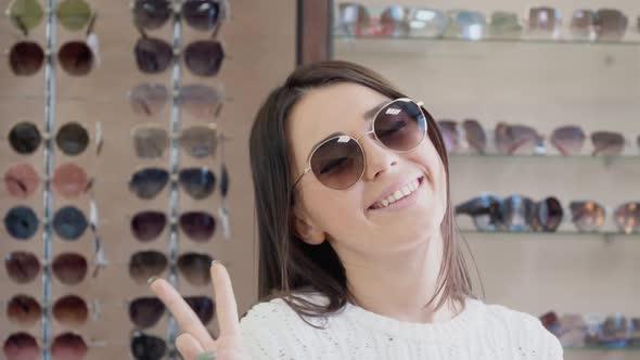 Cheerful Young Woman in Sunglasses Smiling on the Background of a Shelf with Glasses in the Store