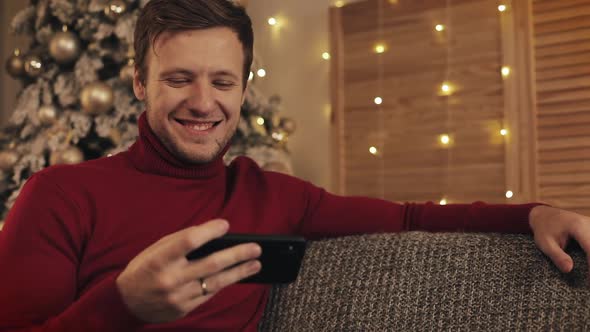 Attractive Man Using Smartphone Sitting on Couch, Messaging, Smiling in Decorated Apartment