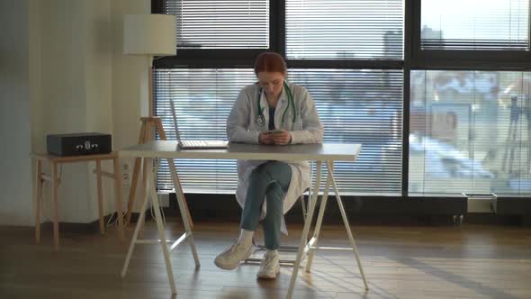 Portrait of Attractive Redhead Female Doctor in White Coat Using Mobile Phone Sitting at Desk with