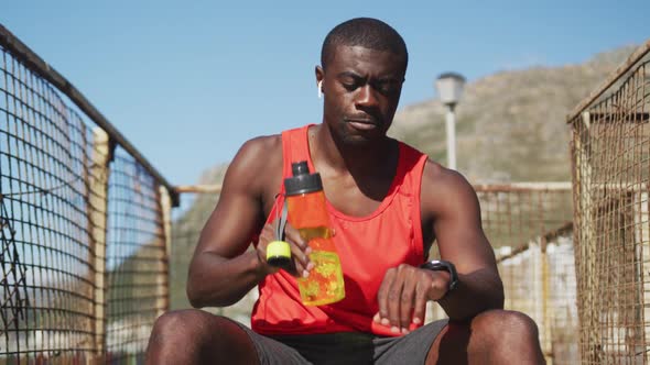 African american man sitting, drinking from water bottle, taking break in exercise outdoors