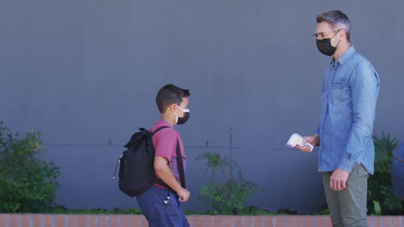 Diverse male teacher measuring temperature of schoolboy, all wearing face masks