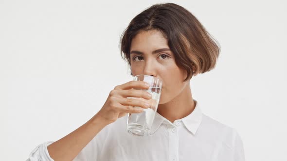 Young Beautiful Brunette East Asian Female in Shirt Drinking Water From Glass on White Background in