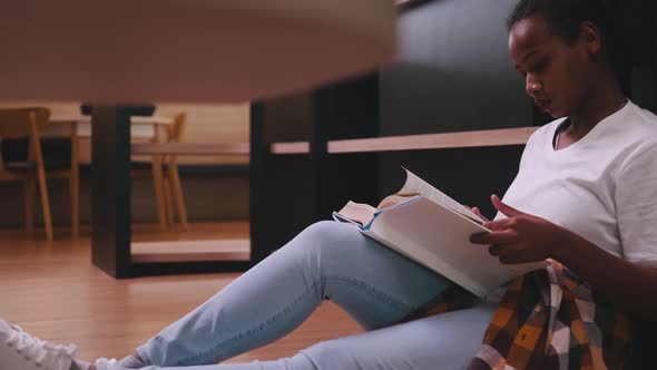 A schoolgirl sitting in front of library's bookshelf and reading a book preparing for an exam.