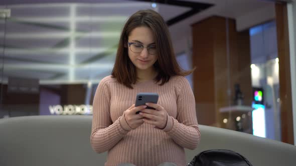 A Young Pregnant Woman in Glasses Sits with a Phone on a Bench