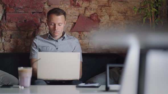 A Young Man in a Shirt is Sitting at a Table with a Laptop and Typing on the Keyboard