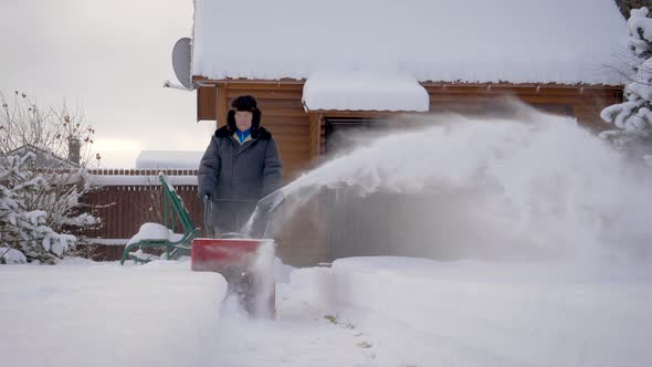 Man Cleans Snow With Snow Plow Background Of Wooden House In Winter