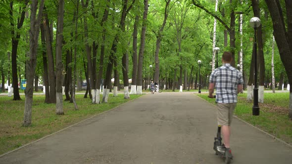 A Young European Man in a Shirt and Shorts Rides an Electric Scooter in the Park