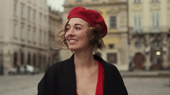 Smiling Young Woman In Red Beret And Red Dress Walking On City Square.