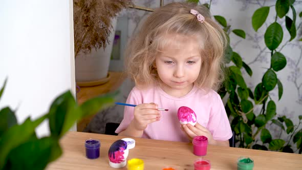 a Little Blonde Girl is Decorating an Easter Egg and Smiling at Home at the Table