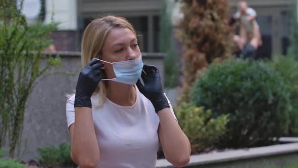 Portrait of Pretty Young Woman in Black Gloves Looking Into the Camera and Putting on Medical Mask