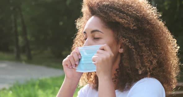 Young Black Woman Putting on a Medical Mask Outside in the Park Sitting on a Bench