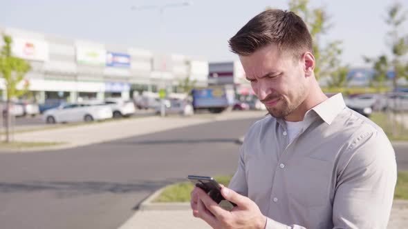 A Young Caucasian Man Works on a Smartphone and Acts Frustrated in an Urban Area