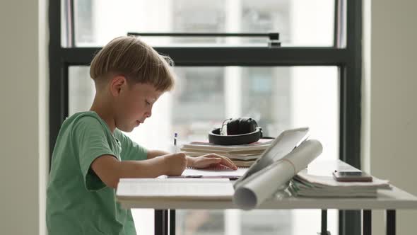 Caucasian Boy Sitting at Table with Workbook and Looking at Tablet Screen While Attentively