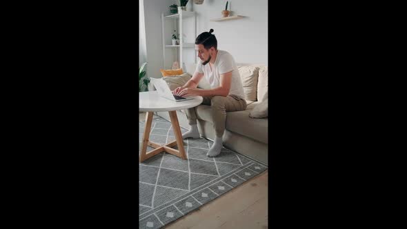 Young Unhappy Man Using Laptop Typing and Working Sitting on the Couch at Home Close Laptop Vertical