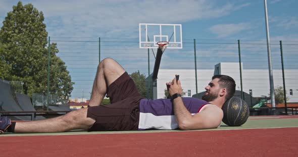 Basketball Player Lying on Floor and Browsing on Smartphone, Outdoor Court Background