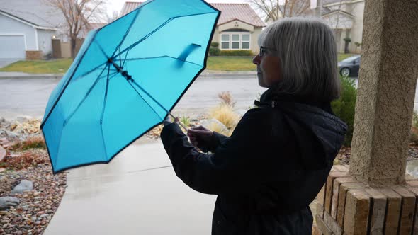 An elderly woman walking out on a wet sidewalk in a rain storm after opening a blue umbrella in slow