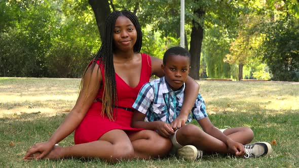 A Young Black Mother and Her Son Sit on Grass in a Park and Smile at the Camera