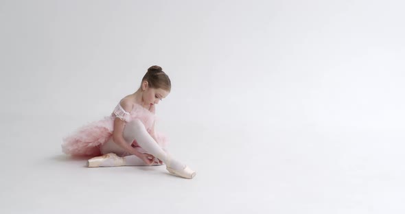 Little Girl in a White Tutu Sits on the Floor and Ties the Ribbons of Her Pointe Shoes Young