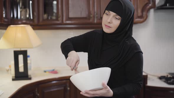 Portrait of Diligent Muslim Woman in Traditional Hijab Standing at Kitchen and Whisking. Young