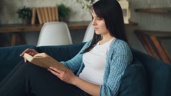 Attractive Young Lady Is Reading Book and Smiling Sitting on Sofa at Home and Relaxing Alone