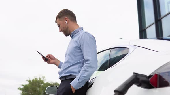 Young Handsome European Man with Wireless Headphones Using Phone While Leaning on His Electric Car