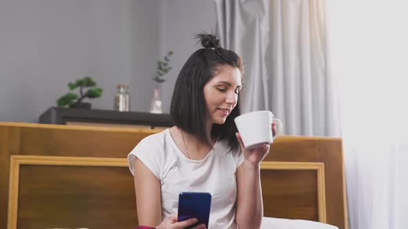 Woman with Dark Hair Watching Funny Video on Phone and Drinking Tea in Bedroom Near Big Window