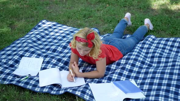 An attractive blond girl is lying on a blanket and draws something in a notebook