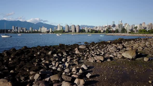 Flying Over The Rocky Shore In Kitsilano Beach With Calm Blue Ocean, Downtown Skyline, And English B